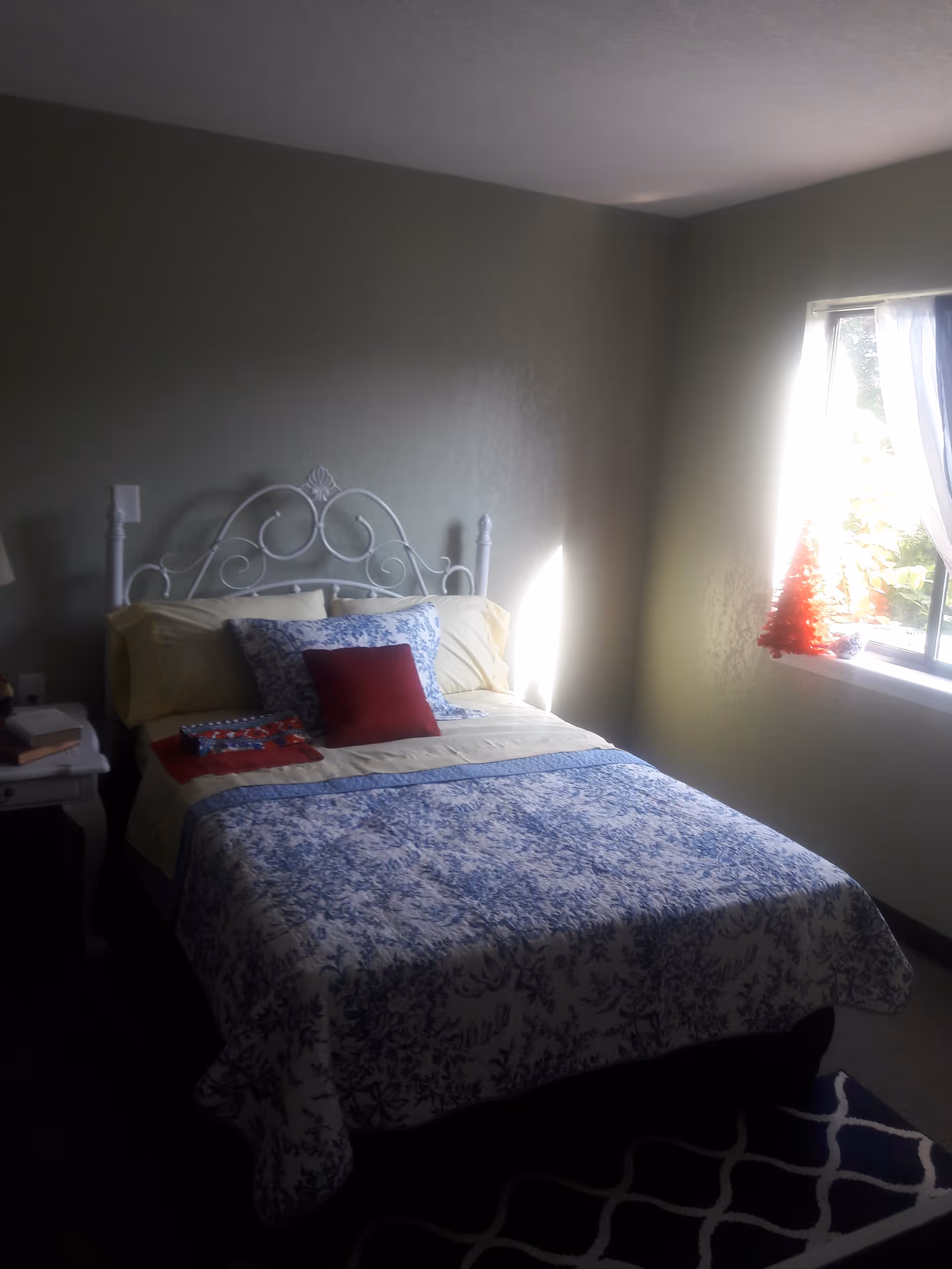 Sunlit bedroom with a white metal headboard and a bed dressed in blue-and-white patterned bedding next to a window with a small red decoration.