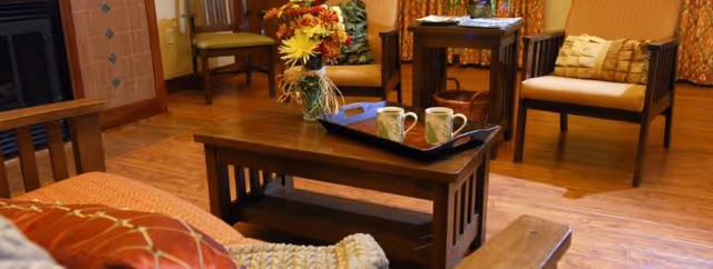 A cozy living room area with wooden furniture including a coffee table with a tray holding two mugs, a sofa with cushions, two armchairs, a side table, and a vase with colorful flowers. The room has warm lighting and wooden flooring.