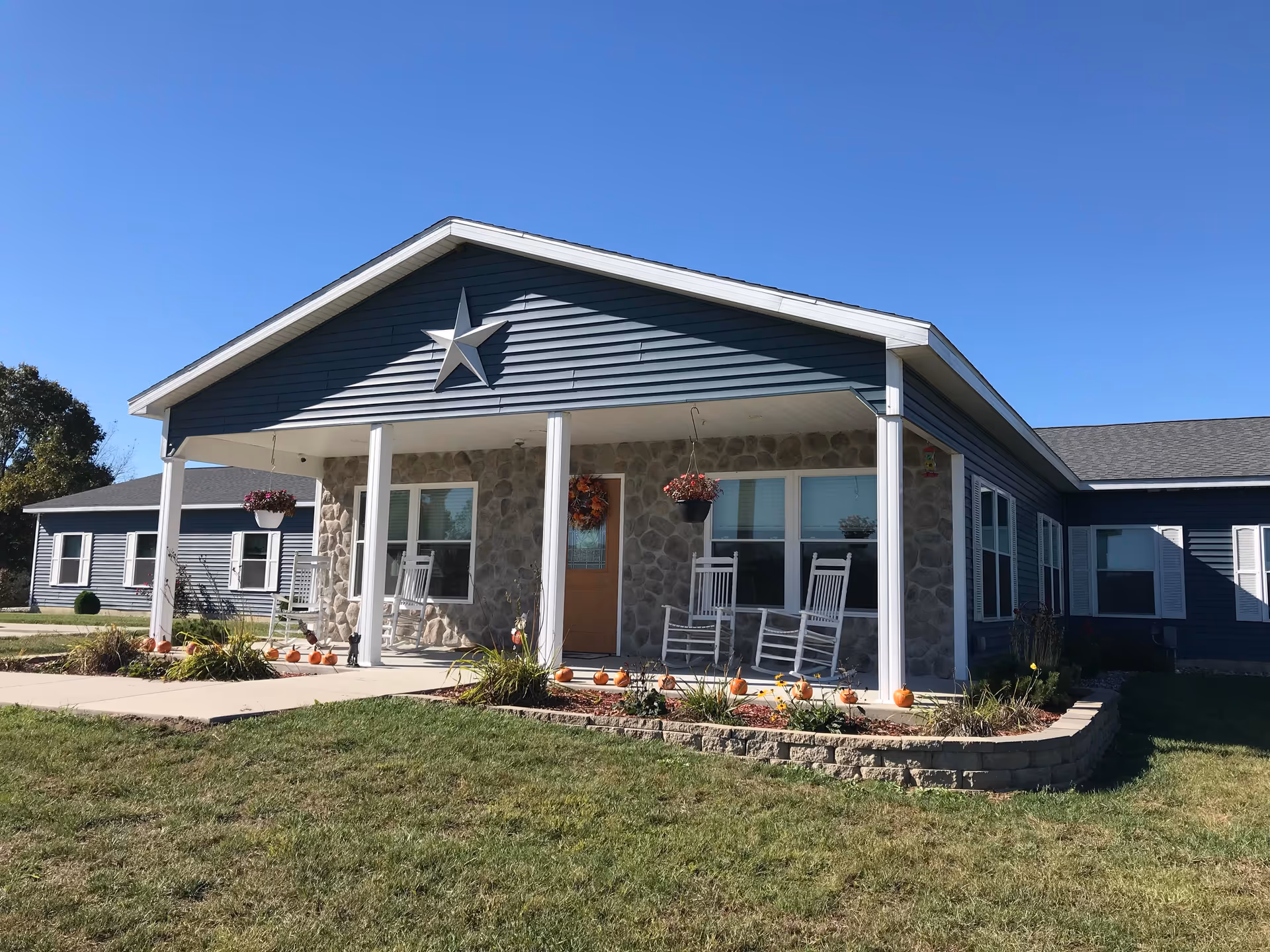 Front exterior view of a single-story assisted living facility with a covered porch featuring white rocking chairs, hanging flower pots, and a decorative star on the gable. The building has blue siding and stone accents around the entrance, with a well-maintained lawn and small pumpkins decorating the flower beds.