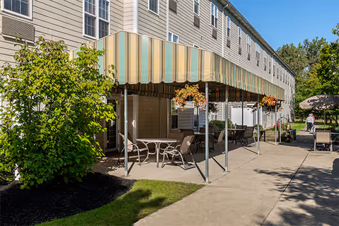 Outdoor patio area at Elderwood Assisted Living at West Seneca with tables and chairs under a striped awning, surrounded by greenery and a concrete walkway. A person is seen walking in the background near a gazebo.