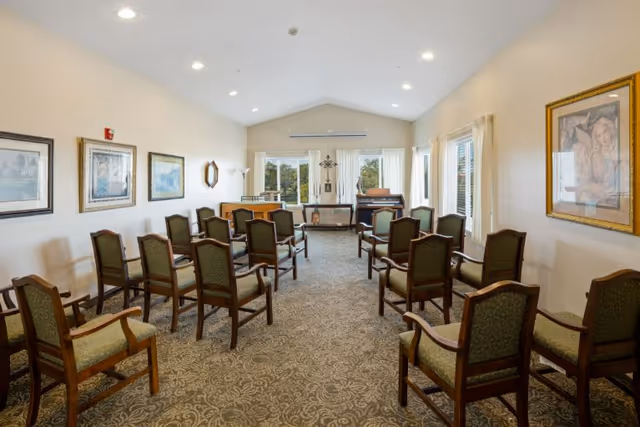 A well-lit room with multiple wooden chairs arranged in rows facing a piano and a small table at the front. The room has beige walls adorned with framed artwork and large windows with white curtains letting in natural light. The carpet has a decorative pattern, and the ceiling is vaulted with recessed lighting.
