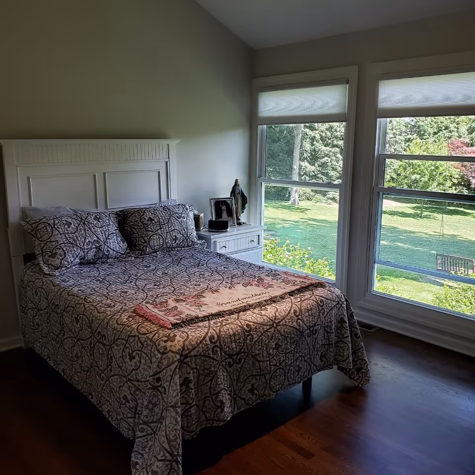Sunlit bedroom with a patterned bed and white headboard beside a nightstand and large windows overlooking a green lawn.
