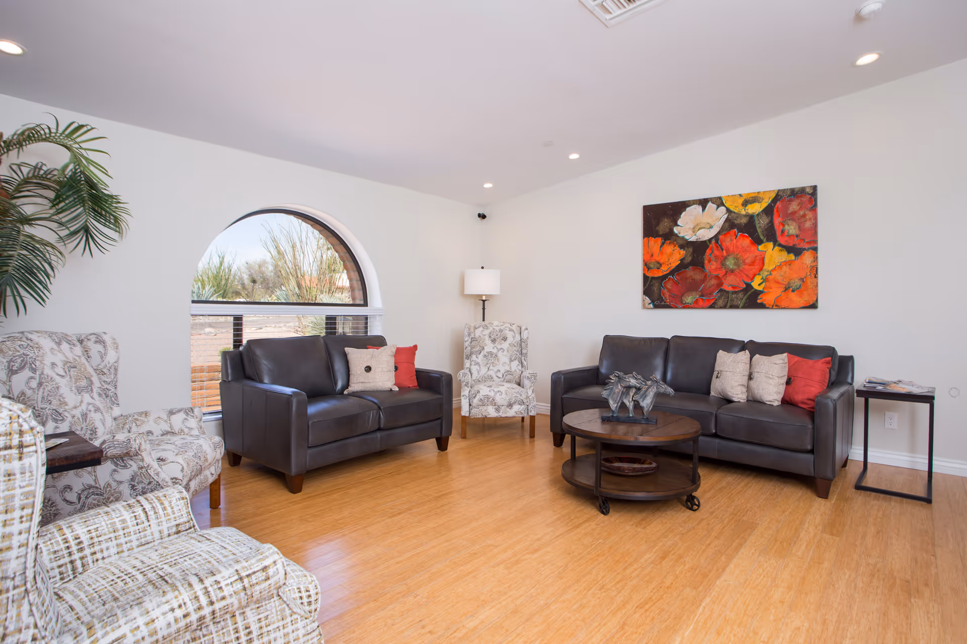 A bright and clean living room with two dark leather sofas, two patterned armchairs, a round wooden coffee table with a horse sculpture, a floor lamp in the corner, and a colorful floral painting on the wall. There is a large arched window showing desert plants outside.