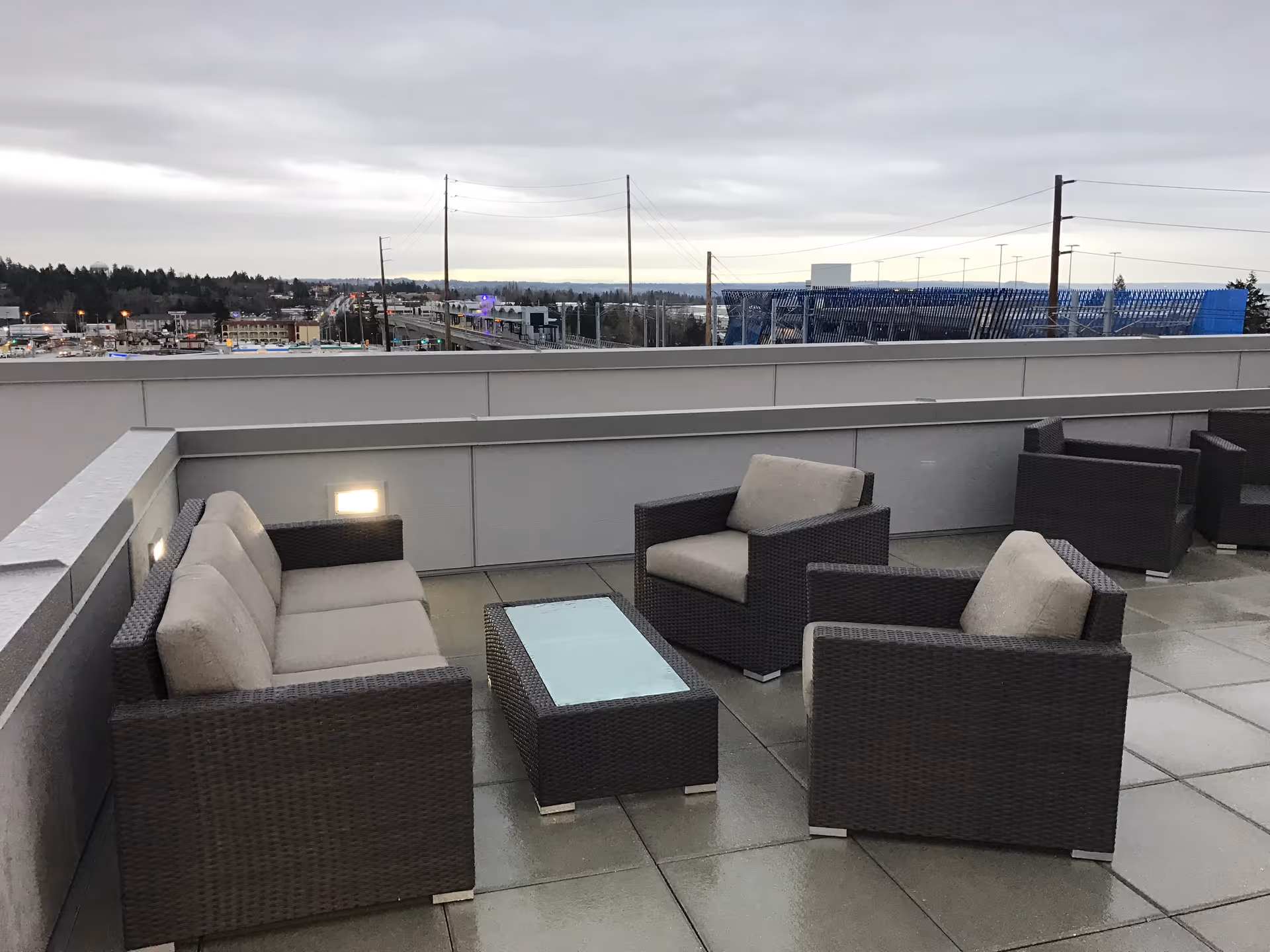 Outdoor patio area with modern wicker furniture including a sofa, two armchairs, and a glass-top coffee table. The patio overlooks a cityscape with buildings, power lines, and a cloudy sky.