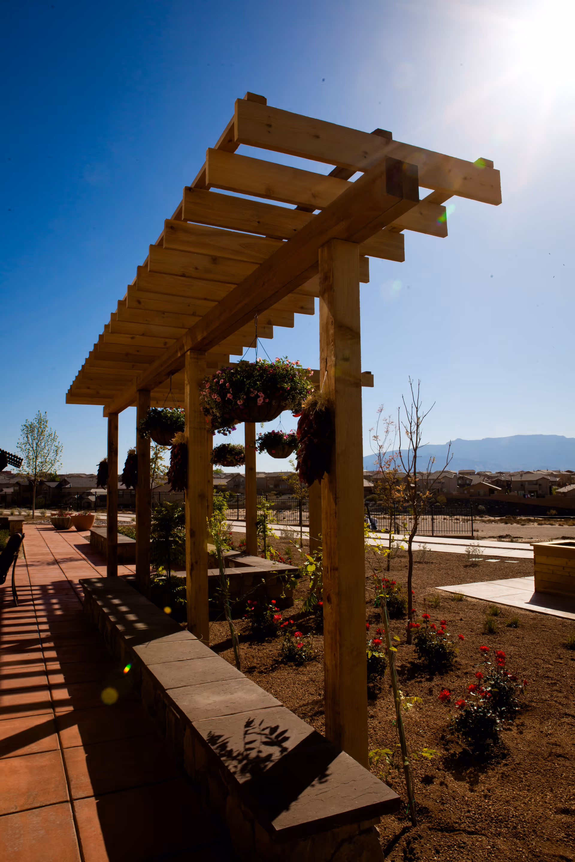 Outdoor patio area with a wooden pergola structure supporting hanging flower baskets. There is a stone bench along the side and a garden with small plants and flowers. The sky is clear and sunny with mountains visible in the background.
