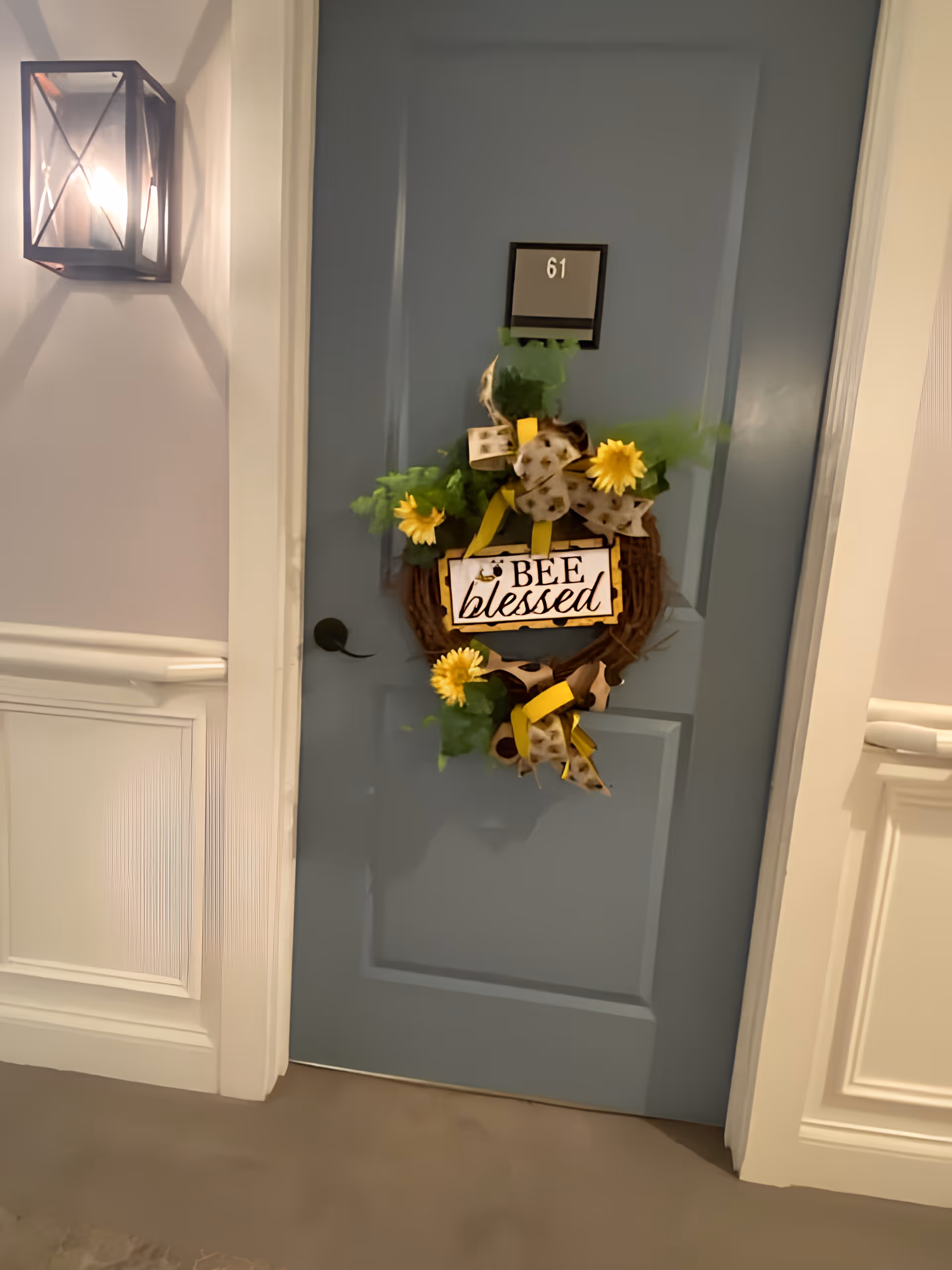 A blue door numbered 61 decorated with a wreath featuring yellow flowers, green leaves, and a sign that reads 'BEE blessed'. The door is set in a hallway with white wainscoting and a wall-mounted lantern-style light fixture on the left.
