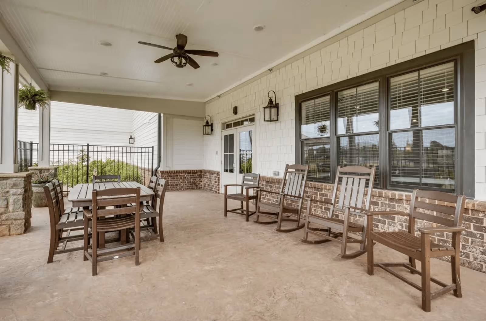 Covered outdoor patio area with a ceiling fan, wooden dining table with six chairs, and a row of wooden rocking chairs and chairs along a brick wall with windows and a door.