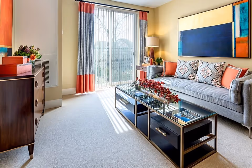 A bright living room with a gray sofa adorned with patterned and solid orange pillows, a glass coffee table with red flowers, a wooden sideboard with decorative boxes and flowers, a floor lamp, and large sliding glass doors with curtains letting in natural light.