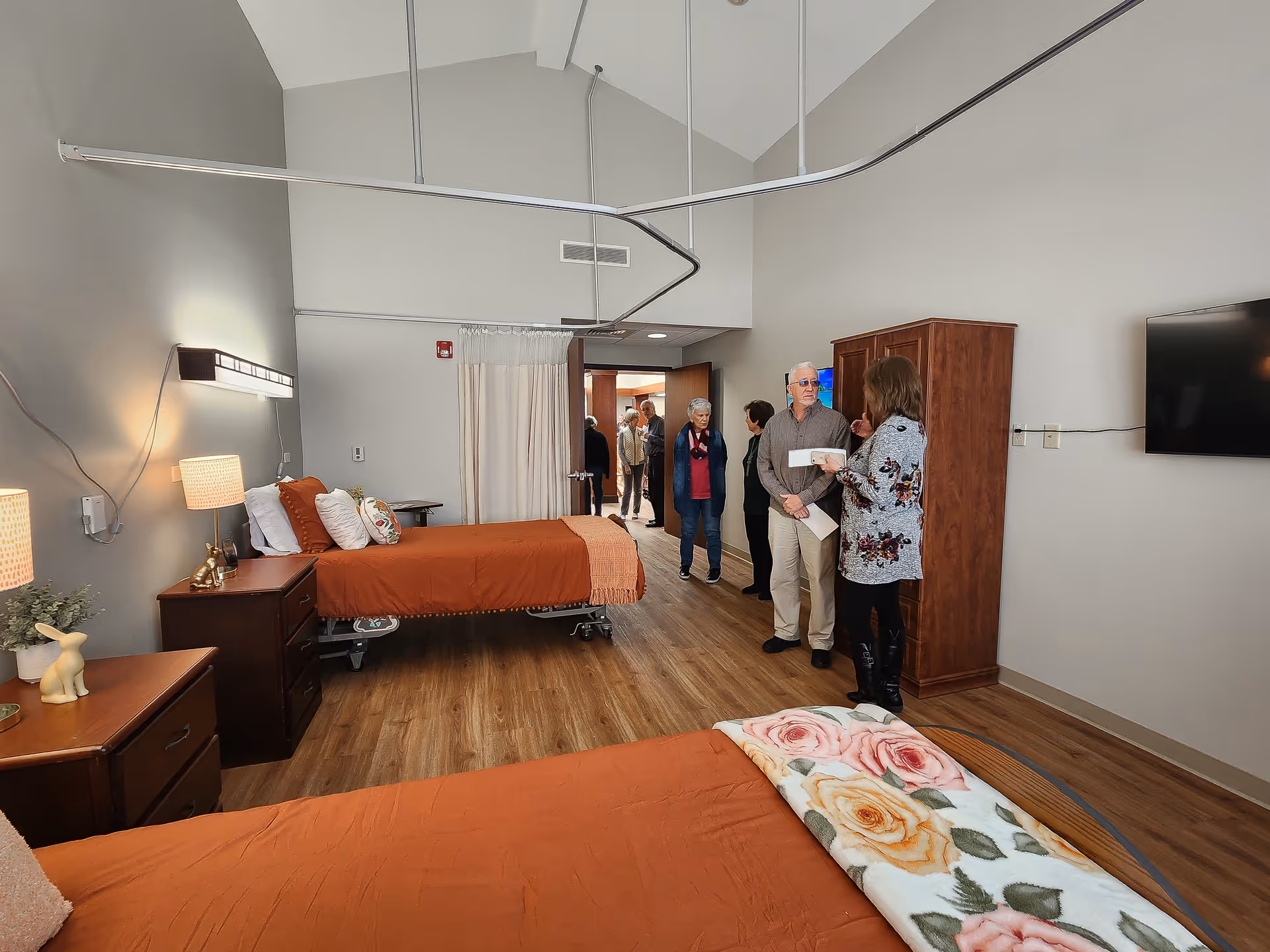 A senior living facility bedroom with two beds covered in orange bedding and floral blankets. There are wooden nightstands with lamps and decorative items. Several people are standing and conversing near the entrance and a wooden wardrobe. The room has wood flooring, high ceilings, and a mounted flat-screen TV on the wall.