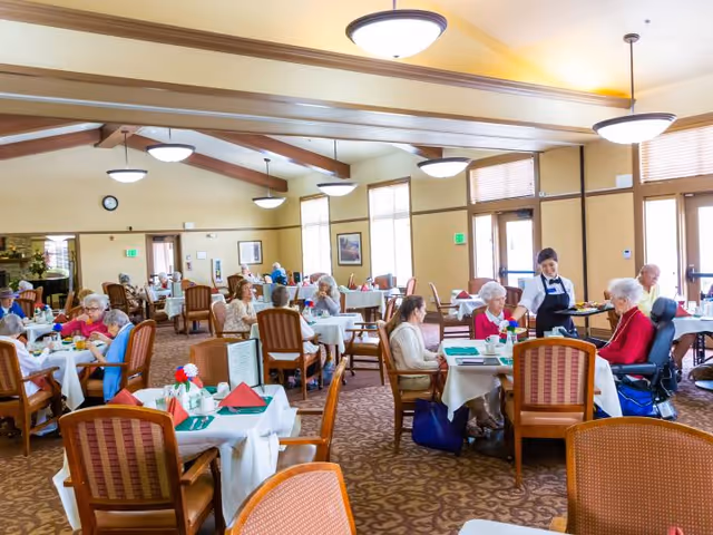 A bright and spacious dining room in a senior living facility with multiple elderly residents seated at tables covered with white tablecloths. A staff member is serving food to one of the residents. The room has large windows, warm lighting fixtures, and wooden chairs with cushions. The atmosphere appears friendly and comfortable.