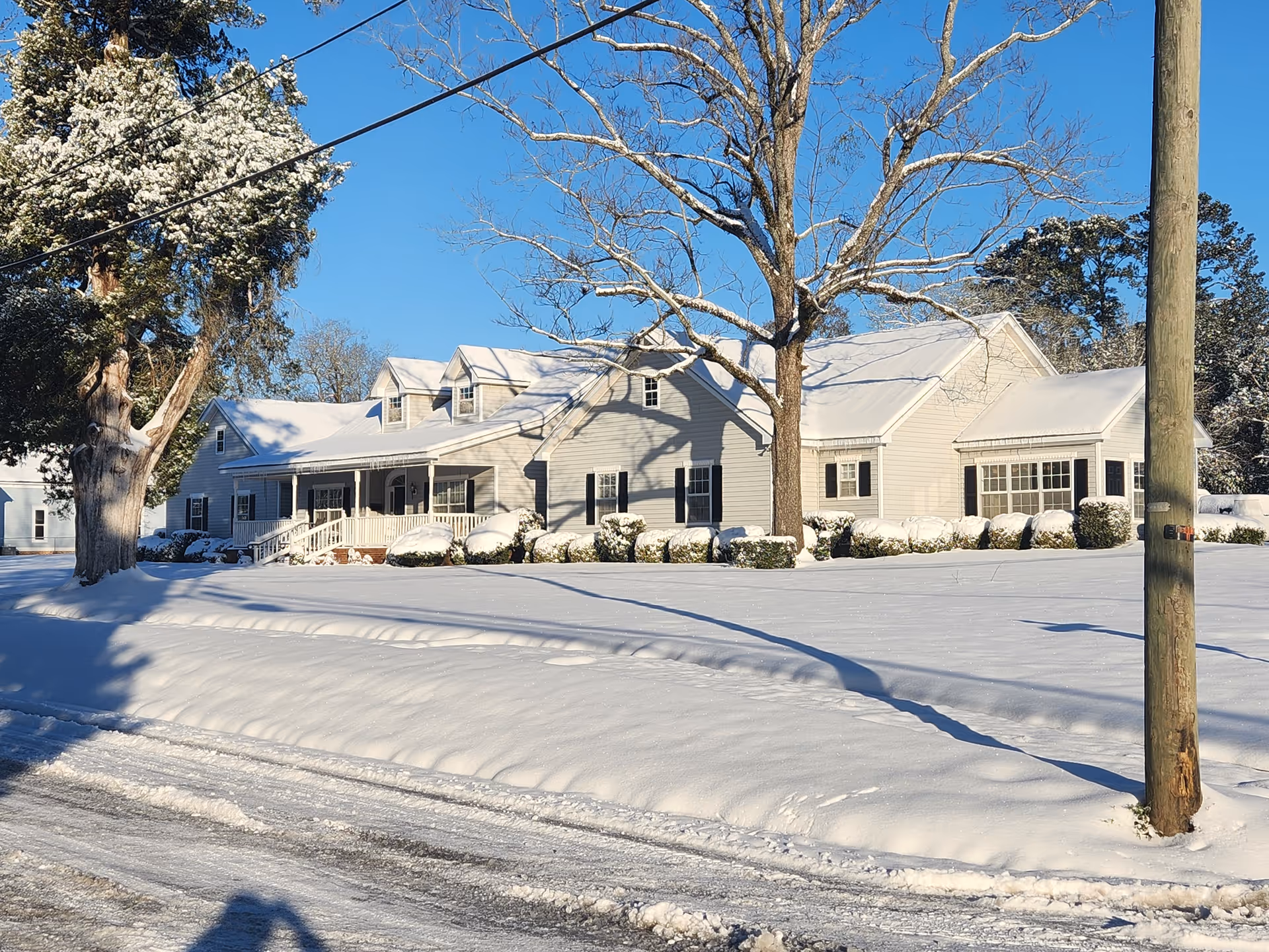 Front view of a snow-covered single-story light-gray building with a porch, leafless trees, and a utility pole under a clear blue sky.