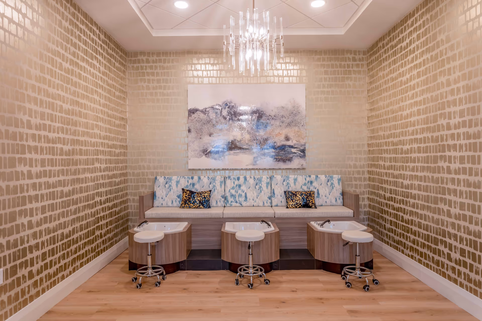A spa pedicure station with three foot baths and stools in front of a cushioned bench, abstract wall art, and a chandelier.