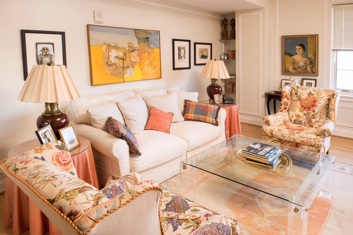 A cozy living room with a white sofa adorned with colorful pillows, two floral upholstered armchairs, a glass coffee table with books on a tray, two side tables with lamps and framed photos, and various framed artworks on the walls.
