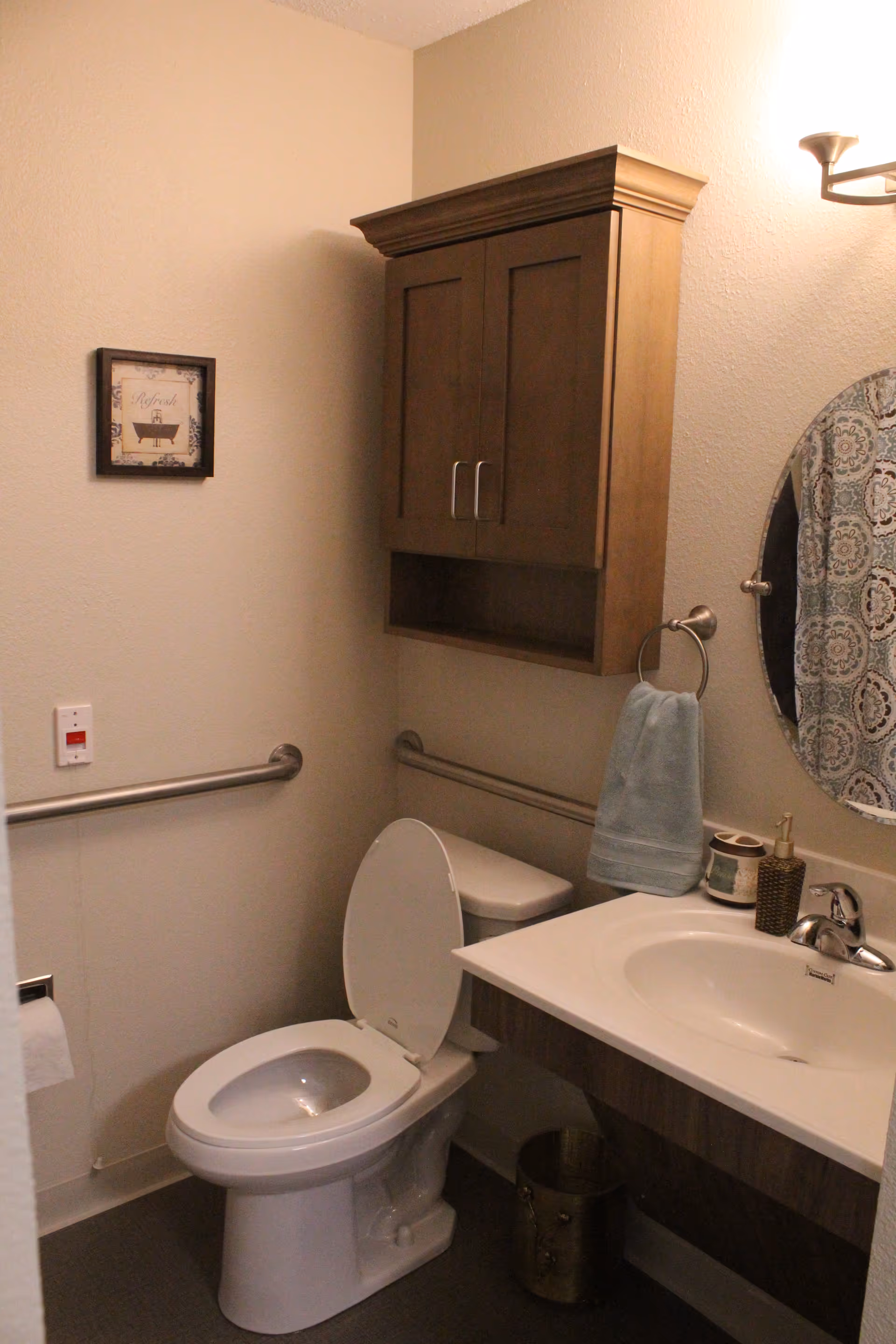 Small accessible bathroom with a toilet (seat up), sink and vanity, wall-mounted cabinet, grab bars, round mirror and towel.