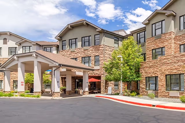 Exterior view of a senior living facility building with stone and beige siding, multiple windows, a covered entrance with pillars, and a tree near the entrance under a partly cloudy sky.