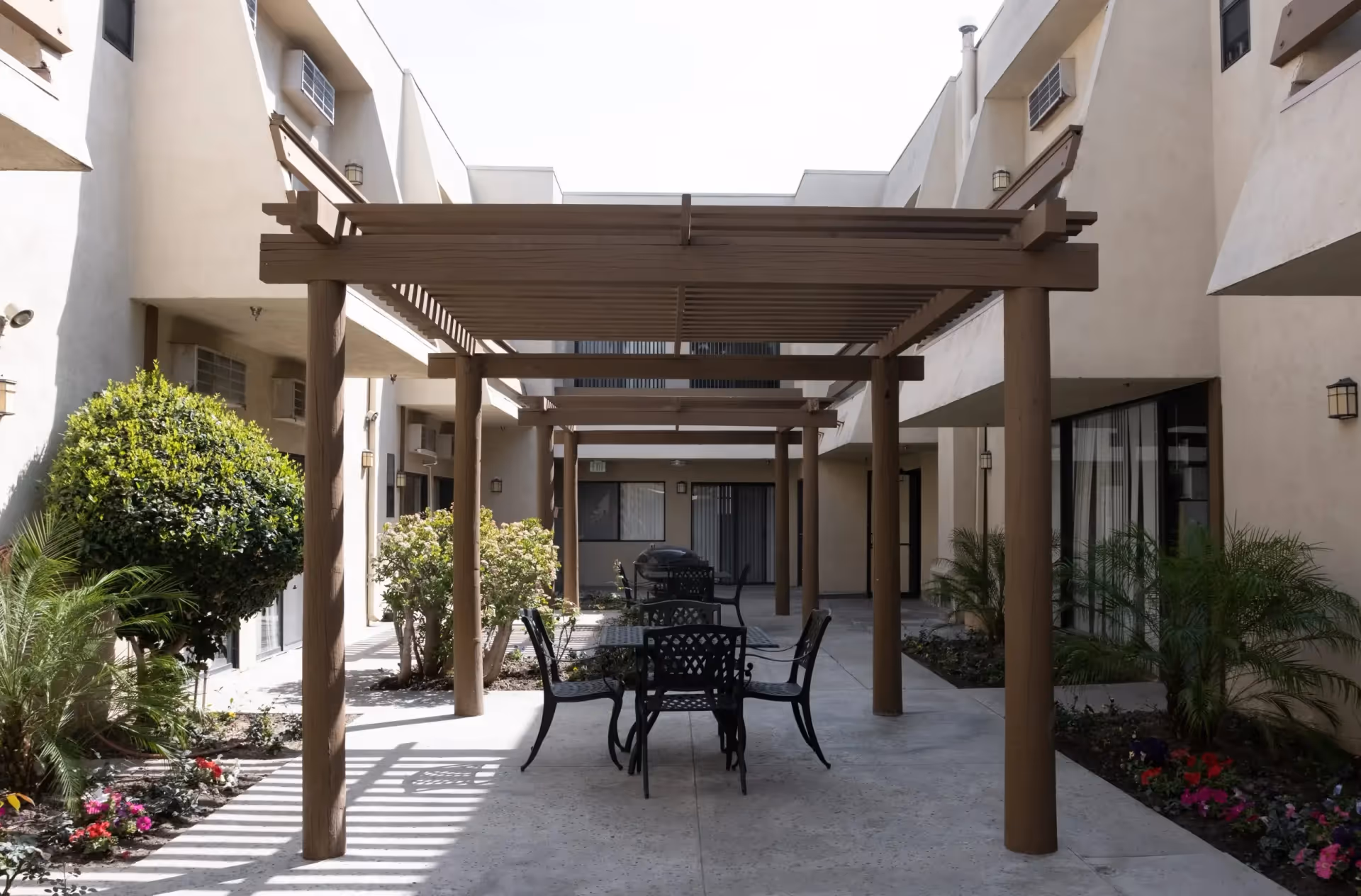 Outdoor courtyard area at Downey Retirement Center featuring a wooden pergola with several black metal tables and chairs underneath. The courtyard is surrounded by beige building walls with windows and doors, and landscaped with green bushes and colorful flowers.