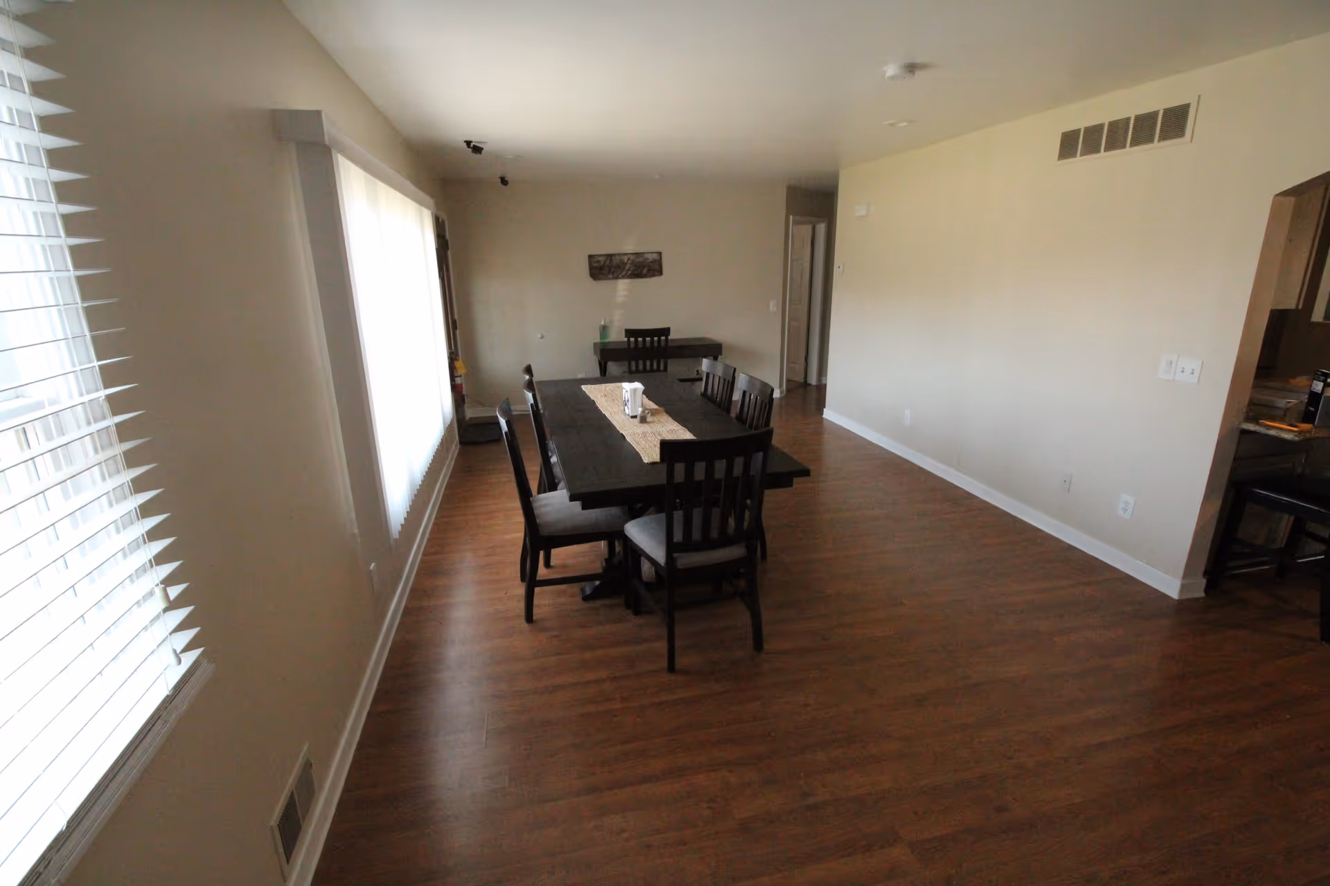 A dining area with a long dark wooden table and six matching chairs on a wooden floor. There is a window with blinds on the left side allowing natural light into the room. The walls are painted beige, and there is a small table with a chair against the far wall. The room has a clean and simple design with minimal decor.