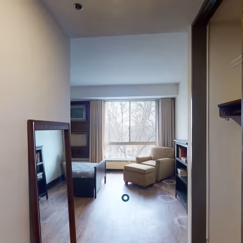 A small furnished bedroom in a senior care facility with bed, chair, mirror, and window.