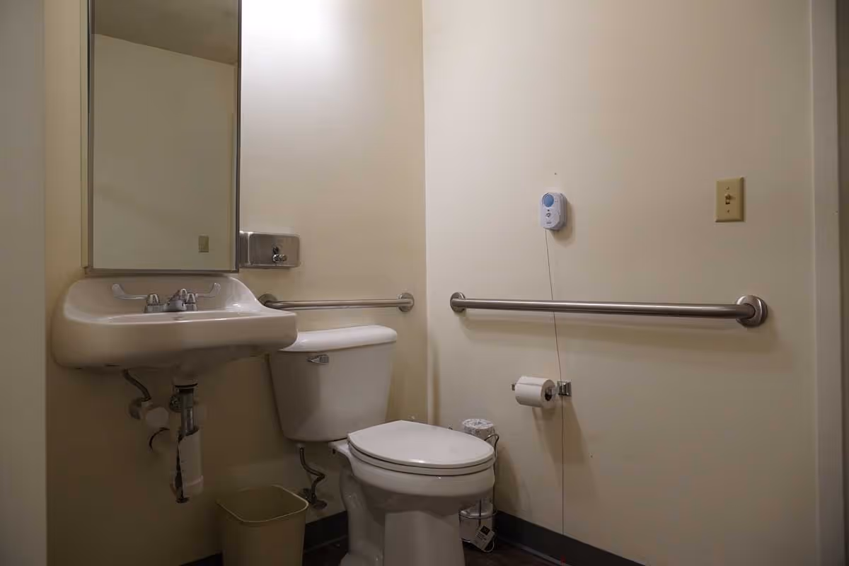 A small bathroom with a white toilet and a wall-mounted sink. There are metal grab bars on the walls next to the toilet and sink, a mirror above the sink, a soap dispenser, a toilet paper holder with a roll of toilet paper, a small trash can, and a wall-mounted emergency call button.