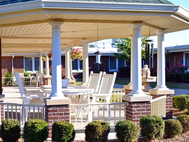 Outdoor covered patio area with white columns and railings, featuring white rocking chairs and tables. Hanging flower baskets and a bird feeder are visible, with a brick building and greenery in the background.