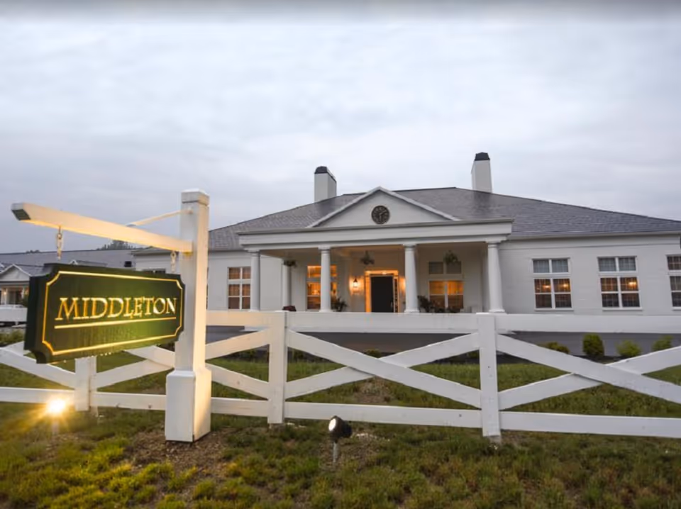 Front exterior of the Middleton Senior Living building at dusk with an illuminated entrance, white fence, and a hanging sign that reads 'MIDDLETON'.