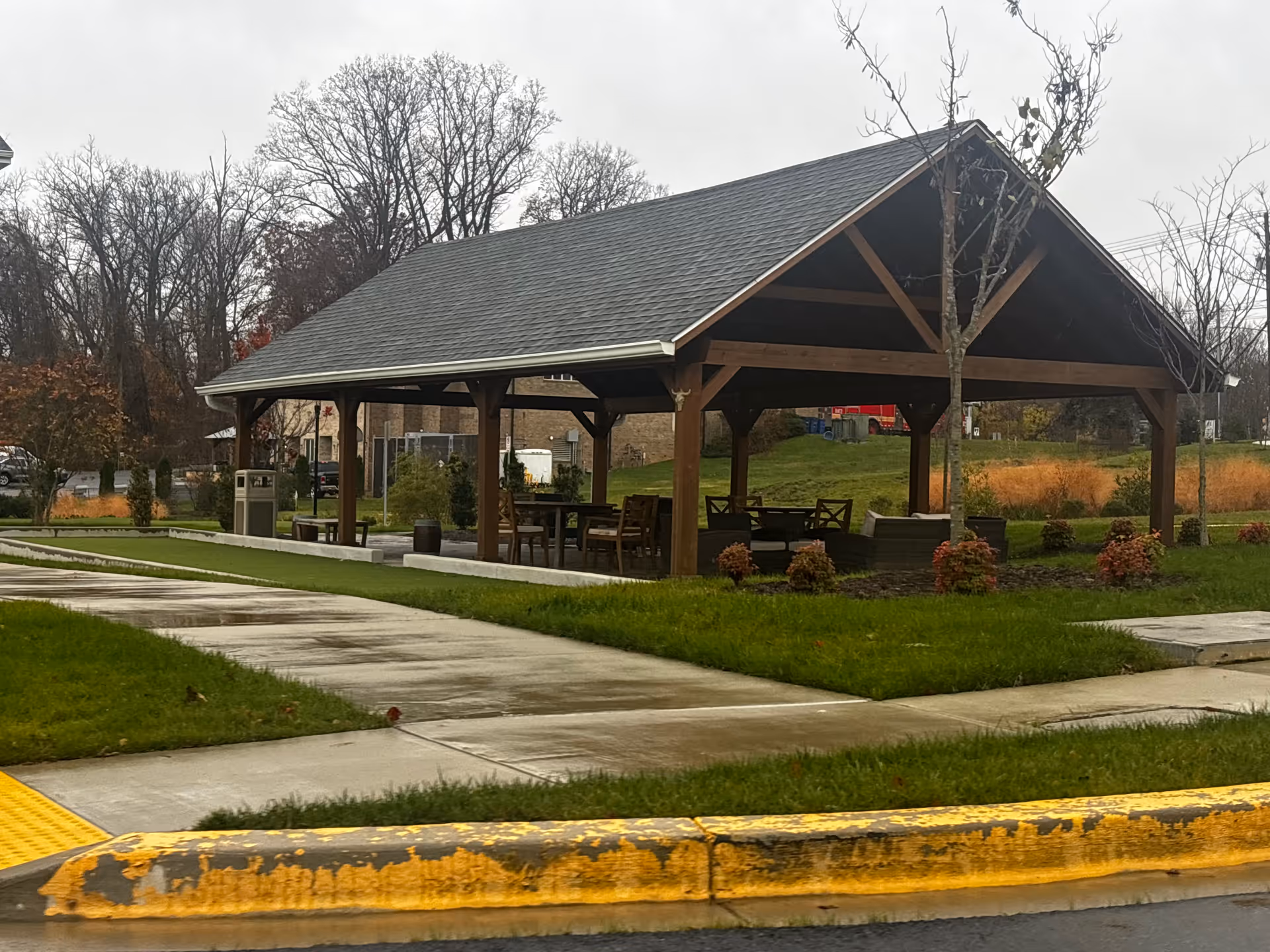 Outdoor covered pavilion with wooden beams and roof, containing tables and chairs, surrounded by green grass, small bushes, and trees on a cloudy day.