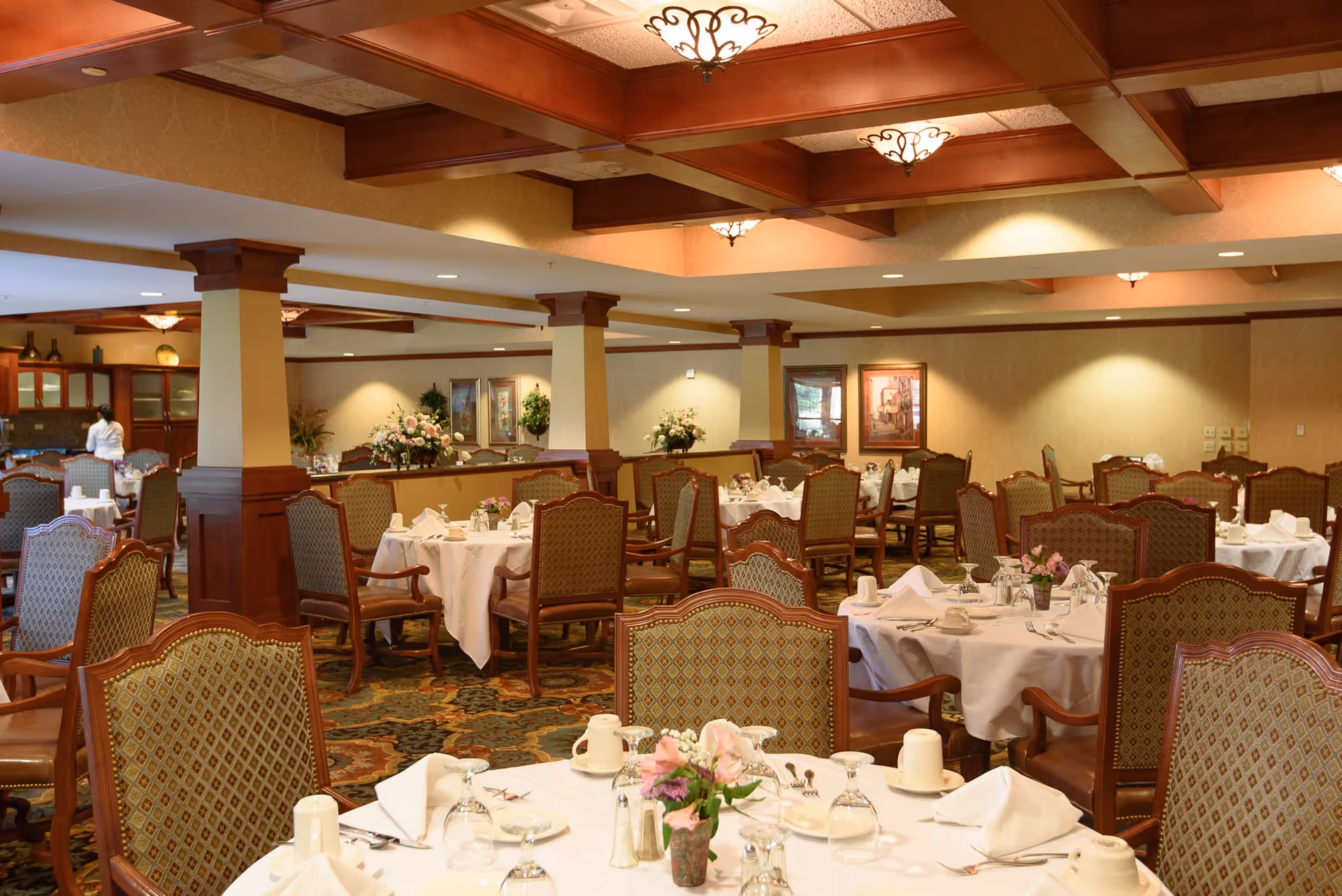 A spacious dining room with multiple round tables covered with white tablecloths, set with plates, glasses, napkins, and silverware. The room features patterned upholstered chairs, wooden columns, decorative ceiling beams, and warm lighting. A person is seen in the background near a cabinet.