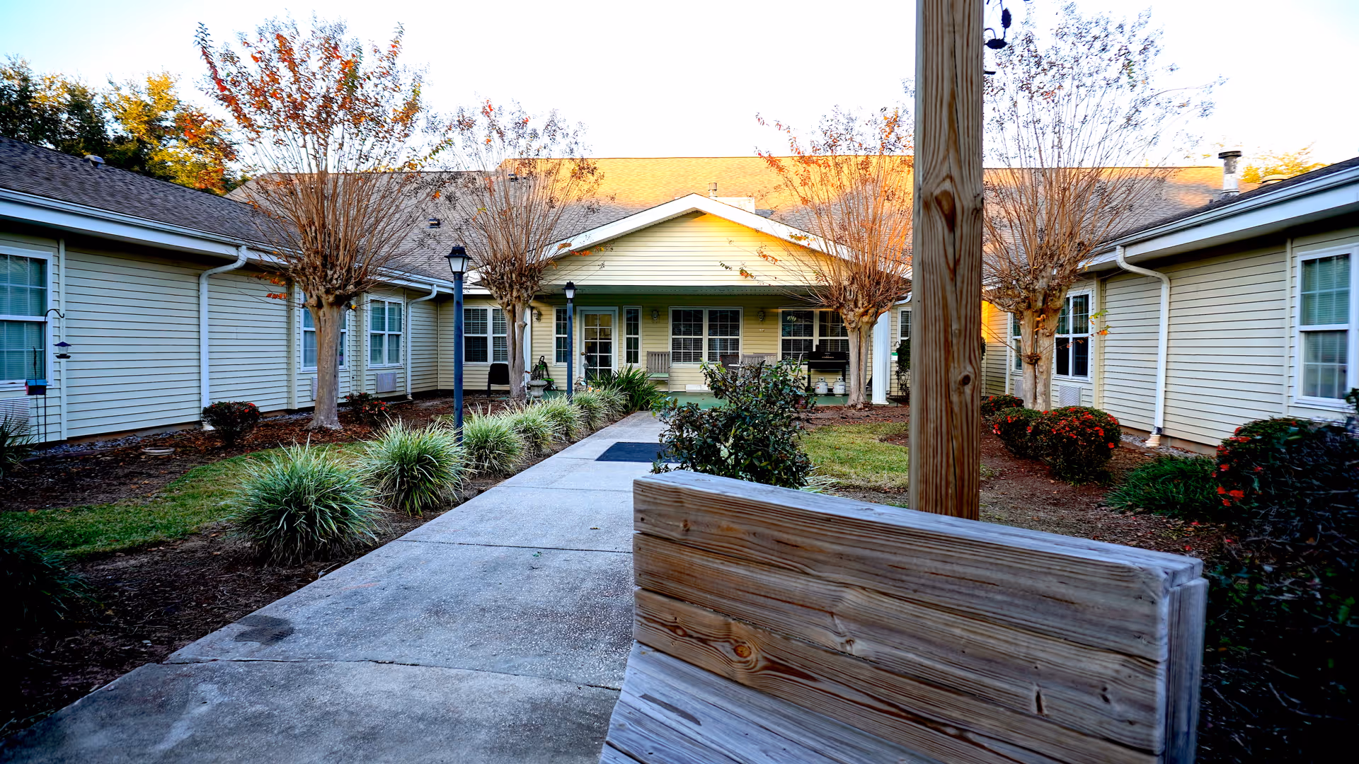 Outdoor courtyard area of a senior living facility with a concrete walkway lined by bushes and small trees, surrounded by single-story beige buildings with white-framed windows. A wooden bench is visible in the foreground.
