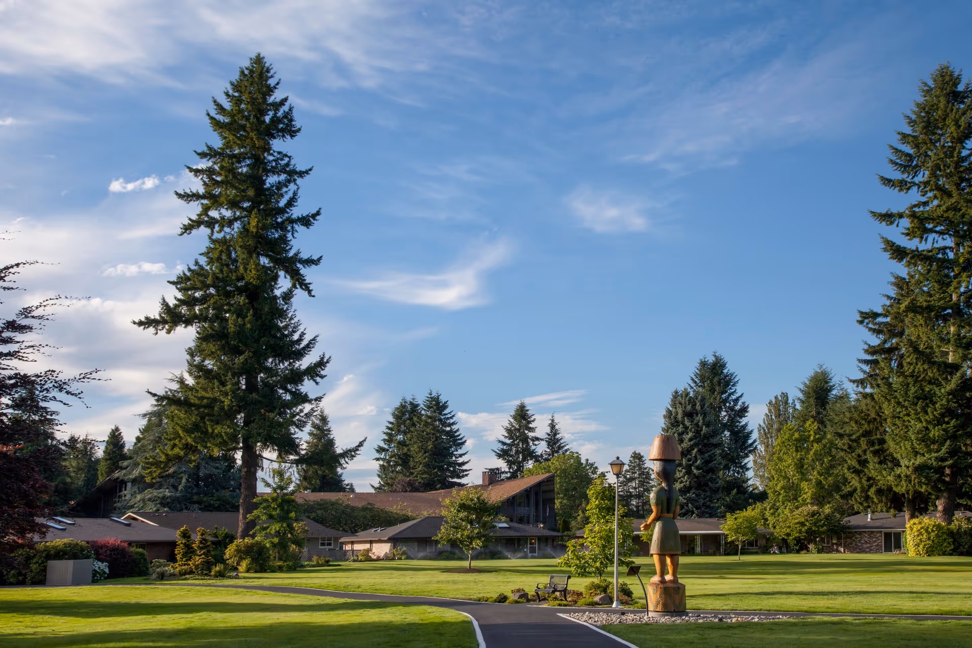 A well-maintained outdoor area of the Panorama facility featuring a paved walkway, green lawns, tall evergreen trees, a wooden statue, and a building with a sloped roof in the background under a partly cloudy blue sky.