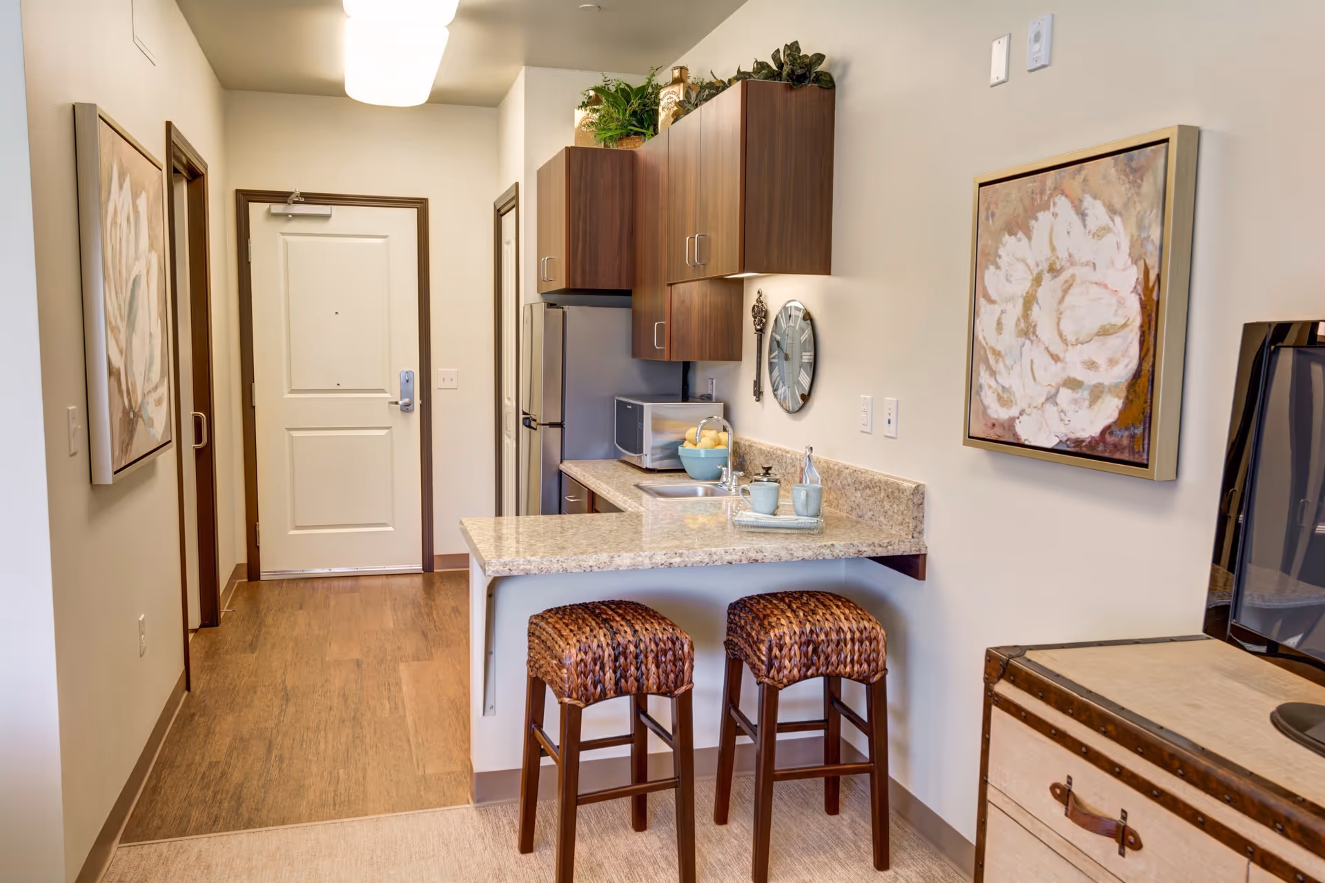 Interior view of a small kitchen area in an assisted living facility. The kitchen features a granite countertop with two woven bar stools, wooden cabinets, a stainless steel refrigerator, a microwave, and a sink. There are decorative plants on top of the cabinets, a wall clock, and framed floral artwork on the walls. The entrance door and hallway are visible in the background.
