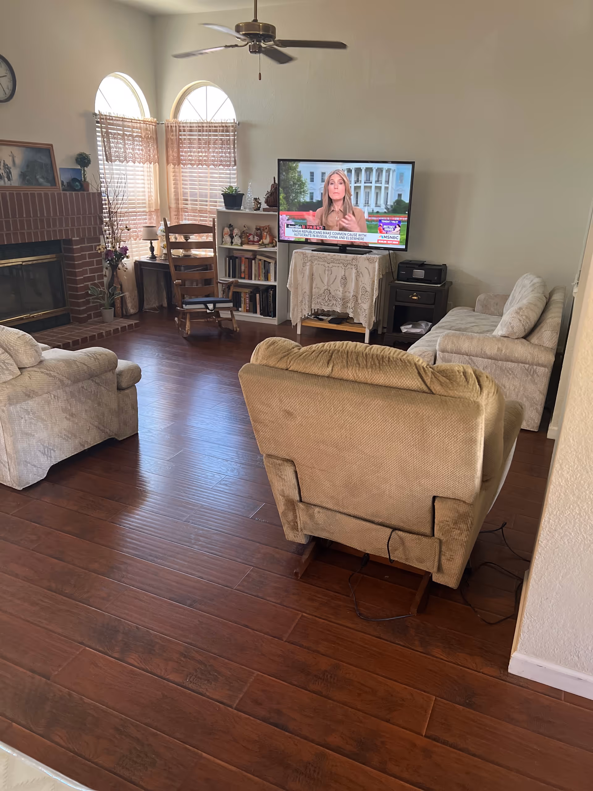 Living room with hardwood floors, a recliner and sofas facing a TV, a brick fireplace, bookshelf, and arched windows.