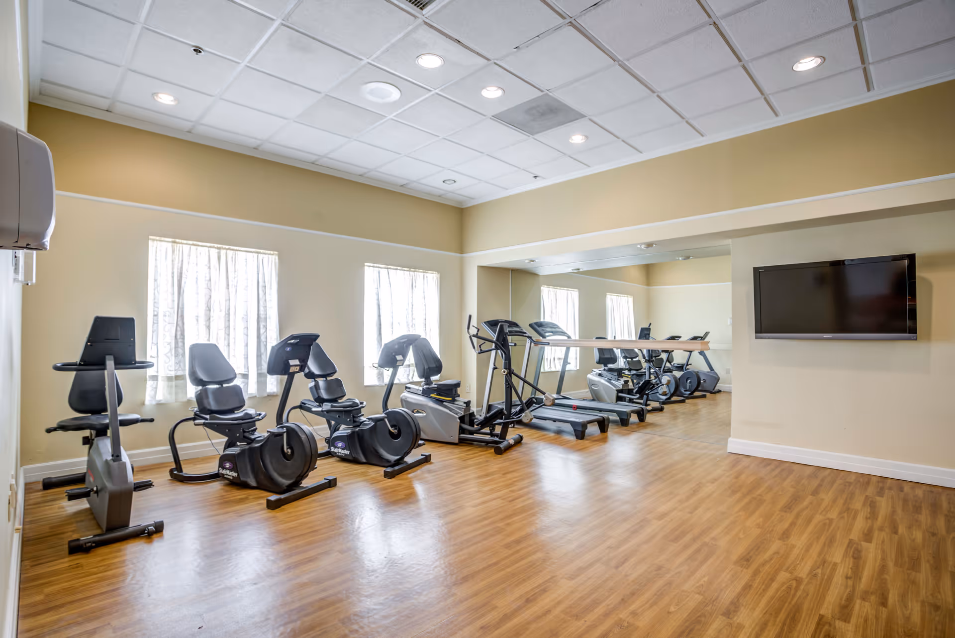 Bright exercise room with recumbent bikes, treadmills, an elliptical machine, wood-look floor, and a wall-mounted TV with mirrors.
