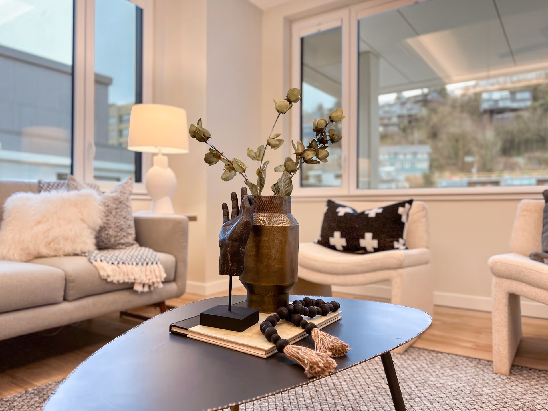 A cozy living room with a gray sofa adorned with fluffy and patterned pillows, a white lamp on a side table, two cream-colored armchairs with black and white patterned pillows, and a black coffee table in the center. On the coffee table, there is a decorative bronze vase with dried flowers, a wooden hand sculpture, and a beaded tassel garland. Large windows in the background show an outdoor view of buildings and trees.