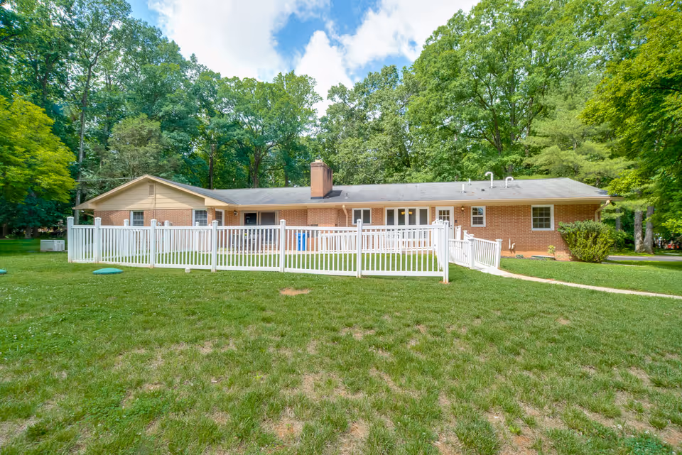 Front view of a single-story brick building with a white fence, grassy yard, and surrounding trees.