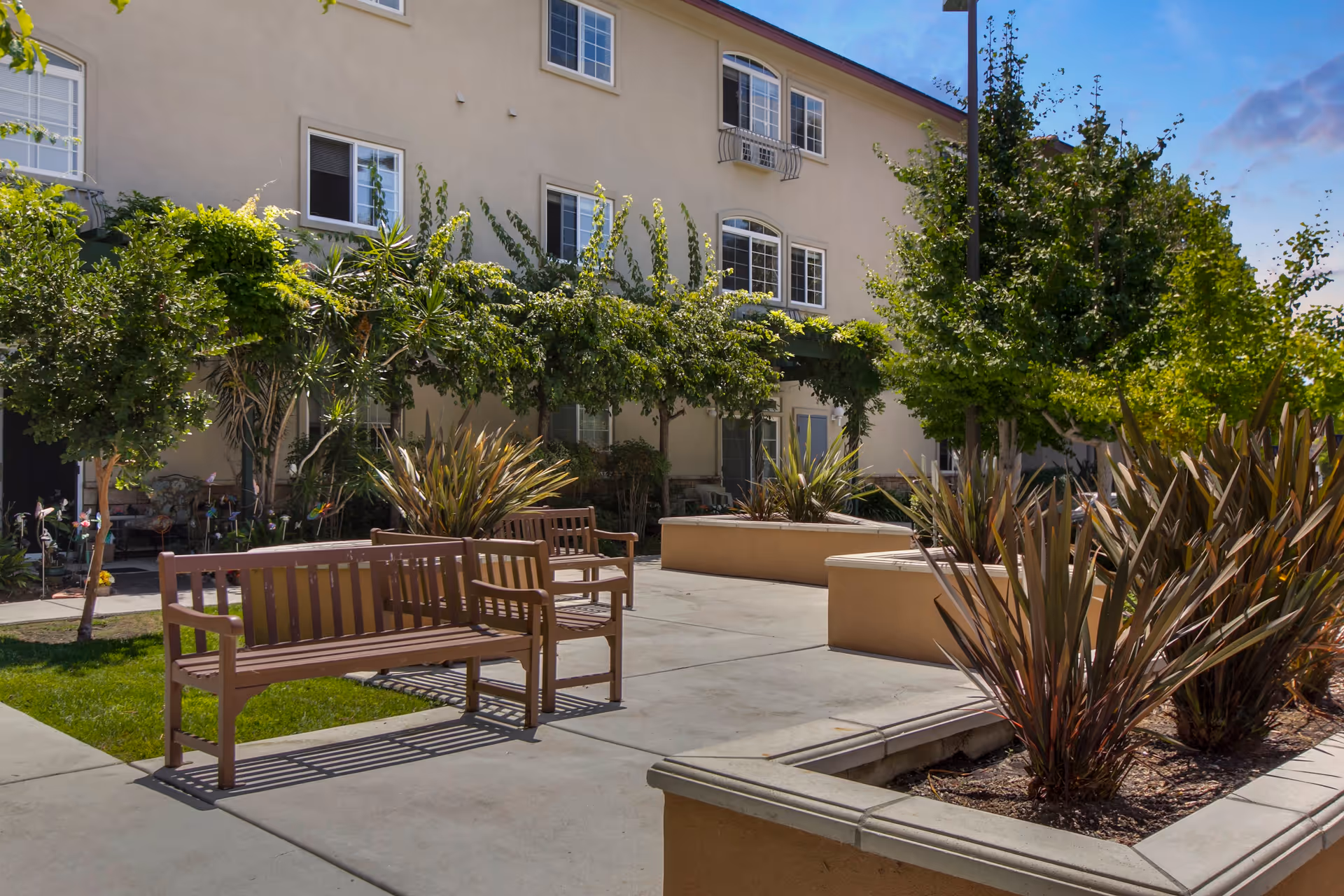 Outdoor courtyard area at a senior living facility with wooden benches, concrete walkways, landscaped plants, trees, and a beige multi-story building in the background under a clear blue sky.