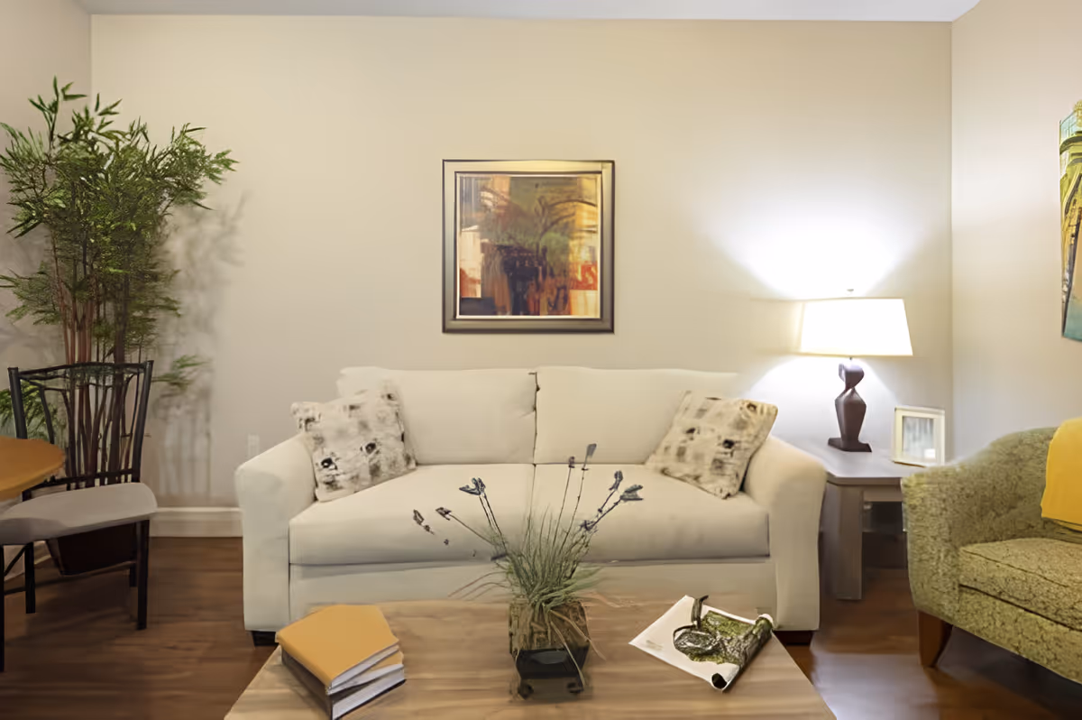 A cozy living room with a white sofa adorned with two patterned pillows, a wooden coffee table with a plant and books, a green armchair with a yellow throw, a side table with a lamp, and framed artwork on the beige walls.