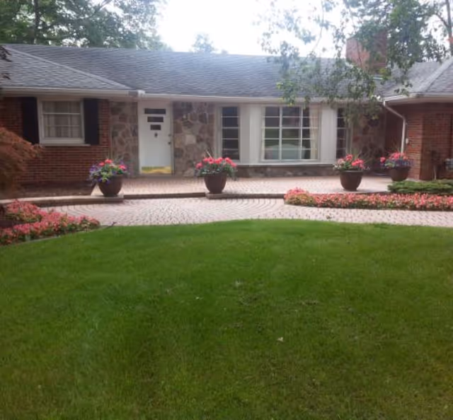 Front exterior view of a single-story brick and stone building with a white door and large windows. There are flower pots with colorful flowers placed on a paved area in front of the building, and a well-maintained green lawn with flower beds in the foreground.