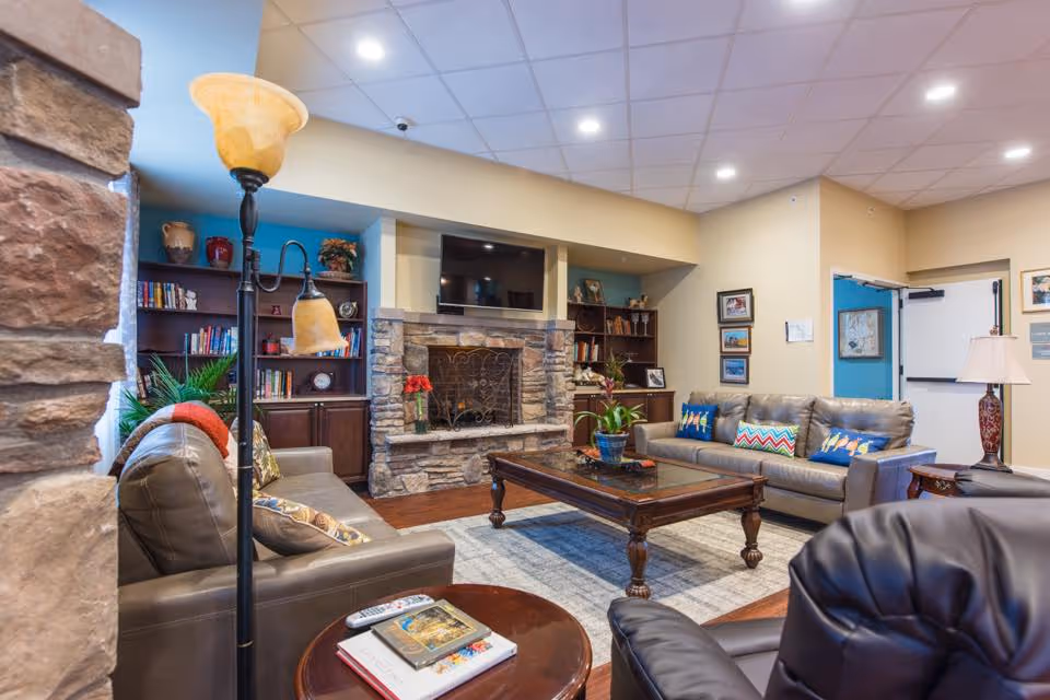 A cozy living room area in a senior living facility featuring two leather sofas with colorful pillows, a stone fireplace with a TV mounted above it, wooden bookshelves filled with books and decorative items, a wooden coffee table with a plant on it, a floor lamp, and framed pictures on the walls.