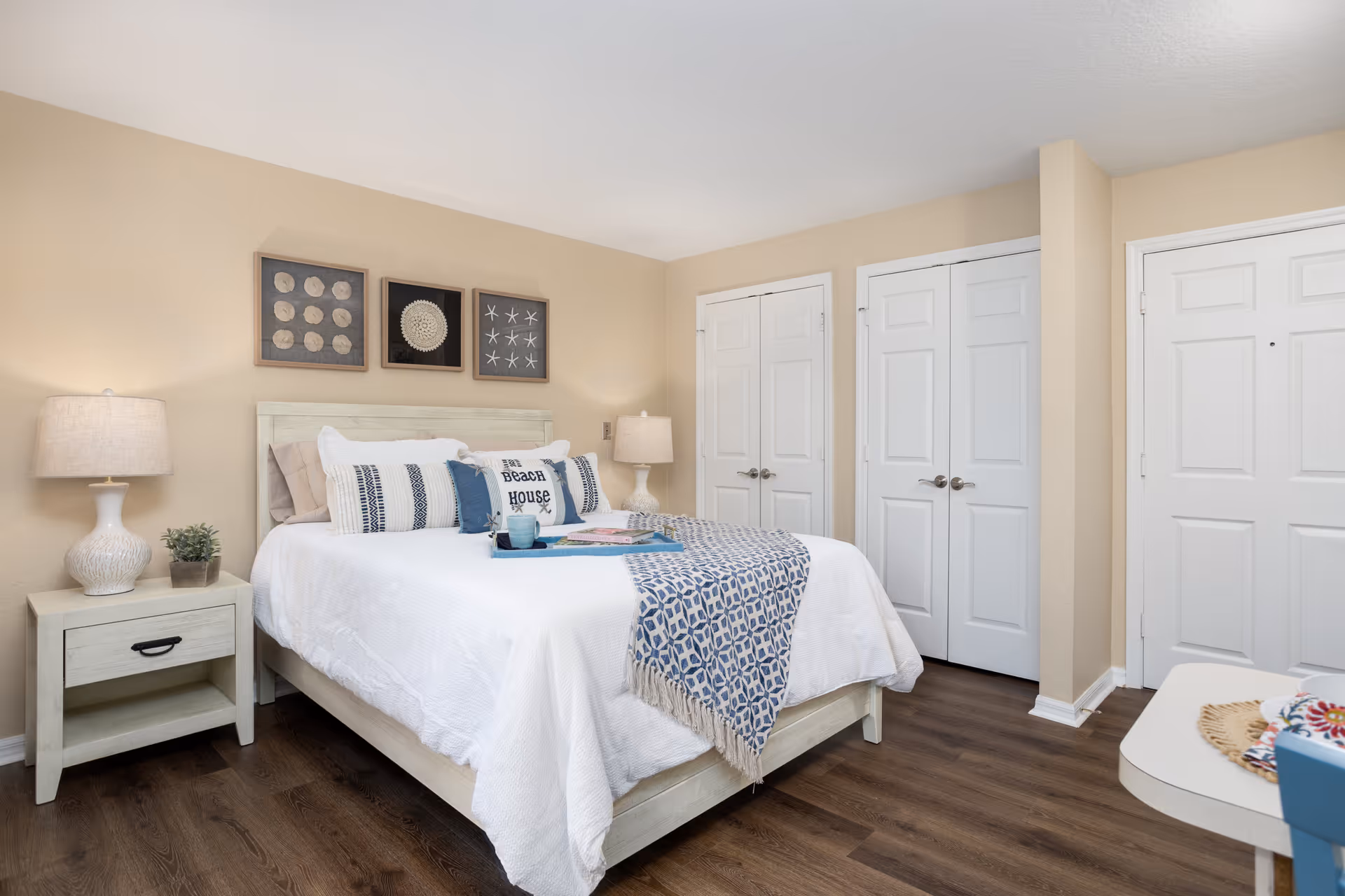 A neatly made bed with white bedding and decorative pillows, including one that says 'Beach House', in a bedroom with beige walls and wooden flooring. There are two lamps on bedside tables, three framed artworks above the bed, and three white doors on the far wall.