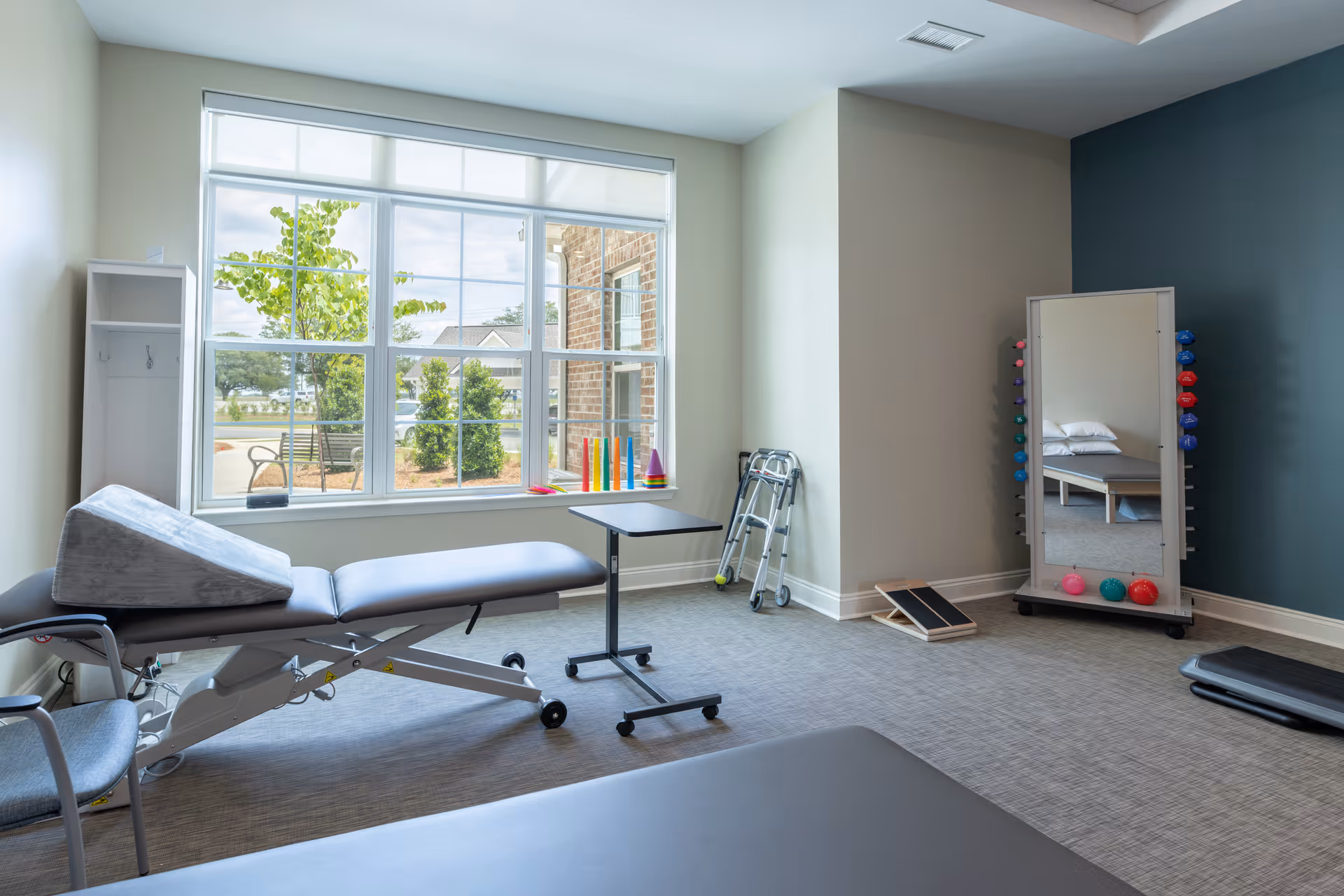 A bright therapy or rehabilitation room with a large window showing outdoor greenery. The room contains a padded therapy table with a wedge cushion, a rolling overbed table, a walker, a mirror with colorful weights attached, and a small ramp. The walls are painted in light neutral colors with one accent wall in blue.
