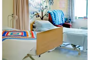 A bright and clean bedroom in a senior living facility with two beds. One bed has a colorful crocheted blanket, and the other has a blue and white patterned blanket. There is a decorative tree mural on the wall behind the beds and a window letting in natural light.