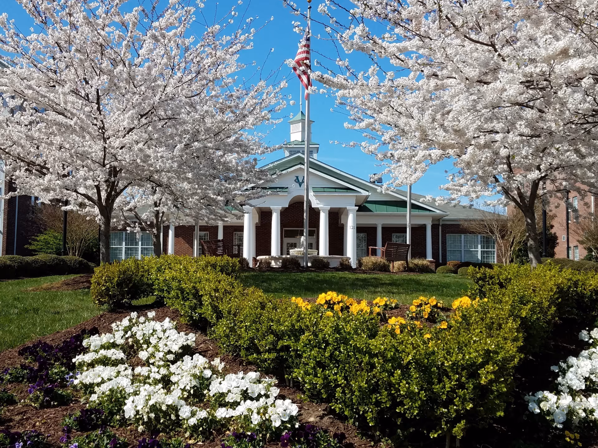 Front view of a brick building with white columns and a green roof, framed by blooming white cherry blossom trees and colorful flower beds under a clear blue sky. An American flag is flying on a flagpole in front of the building.
