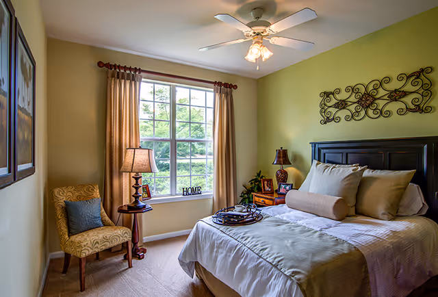 Cozy, well-lit bedroom with a made bed, nightstands, a large window with curtains, an upholstered chair, and a ceiling fan.