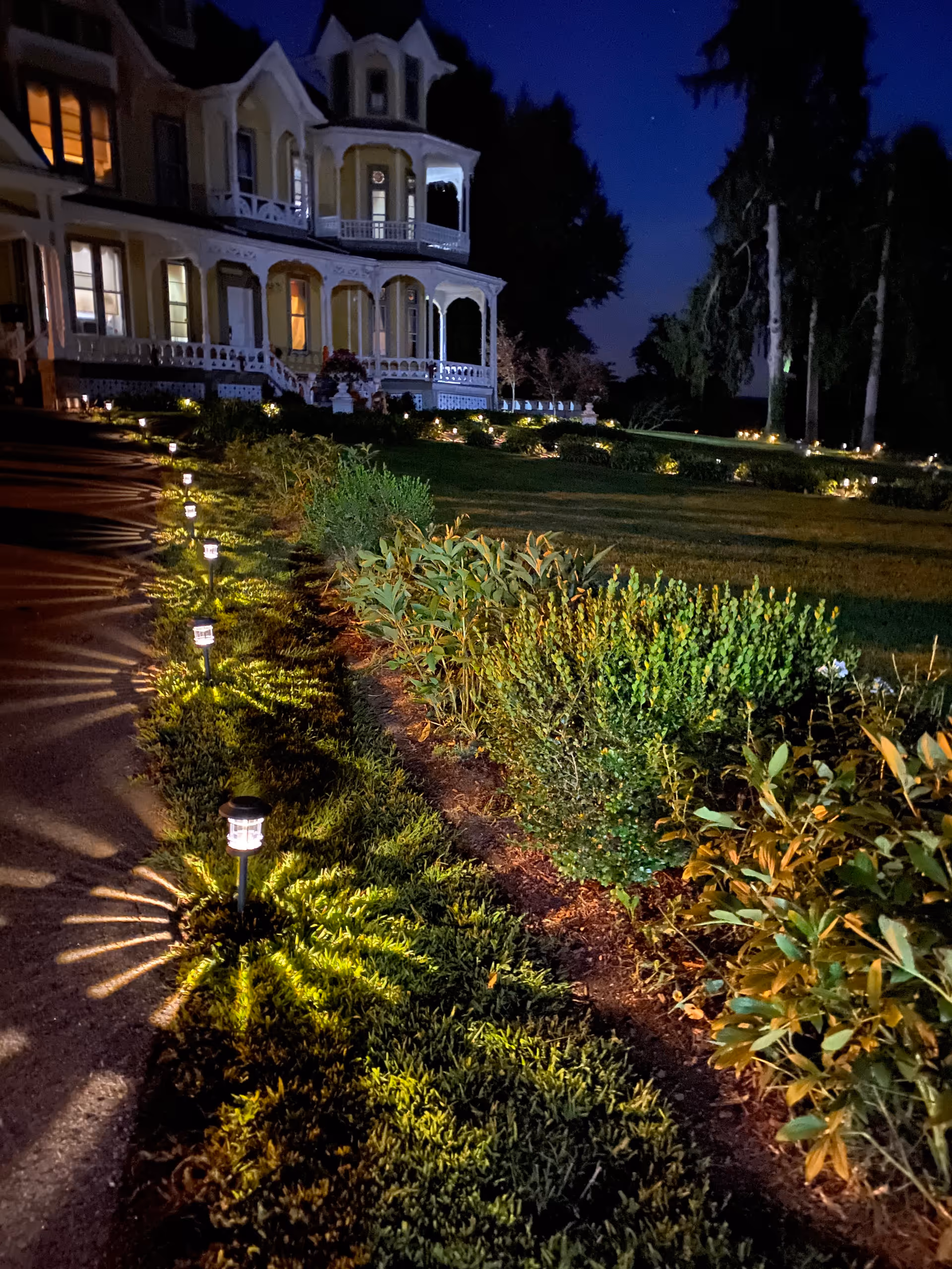 Nighttime view of a large, Victorian-style house with a wraparound porch, illuminated by warm indoor lights. The foreground features a garden with bushes and a pathway lined with small solar lights casting decorative shadows on the grass.