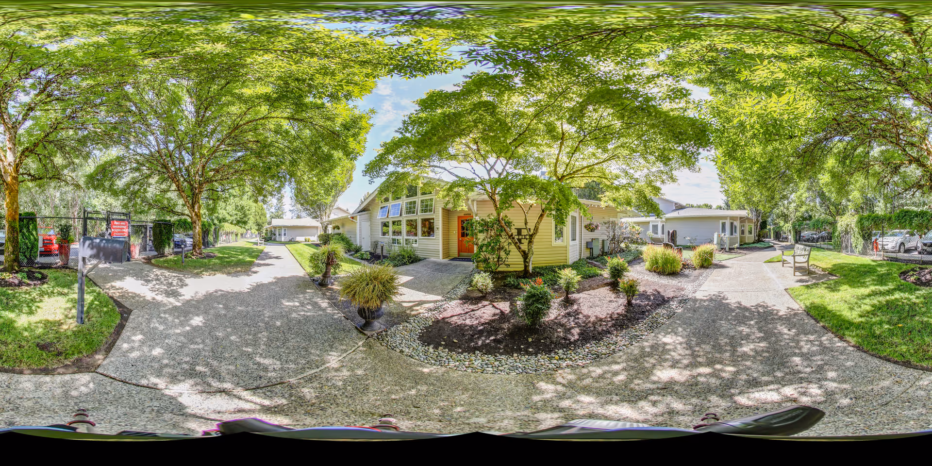 A sunny outdoor pathway at Aegis Living Issaquah, surrounded by lush green trees and landscaped garden beds with shrubs and plants. Several single-story buildings with large windows and light-colored siding are visible along the path. Benches and a parking area with cars can be seen in the background under the shade of the trees.