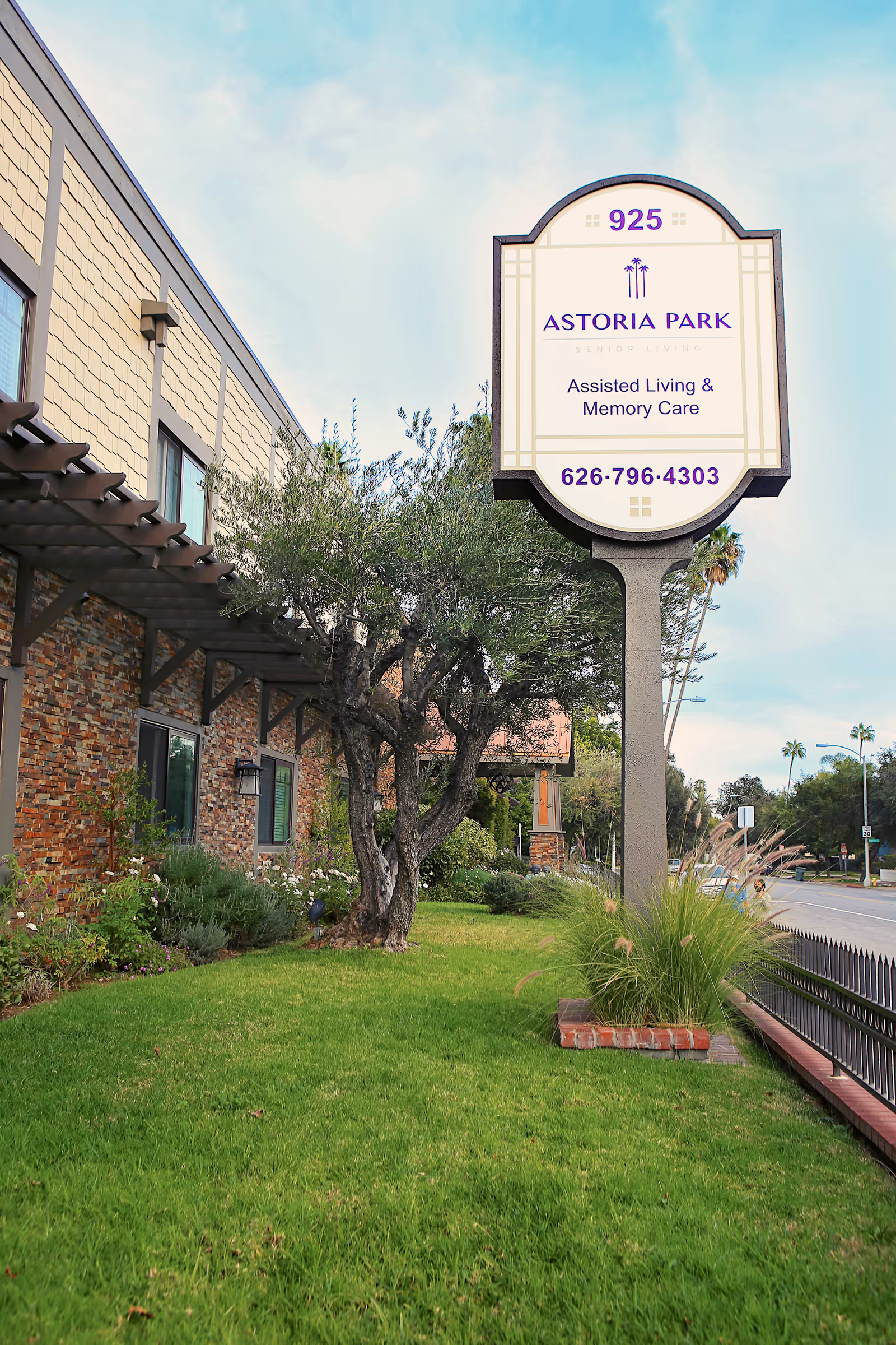 Outdoor view of Astoria Park Senior Living facility showing a large sign with the address 925, the name Astoria Park, and information about assisted living and memory care. The building has a brick and beige exterior with windows, a tree, green grass, and a sidewalk along the street.