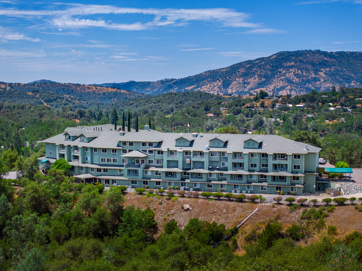A large multi-story senior living facility building painted in light green, surrounded by trees and greenery, with hills and a partly cloudy sky in the background.