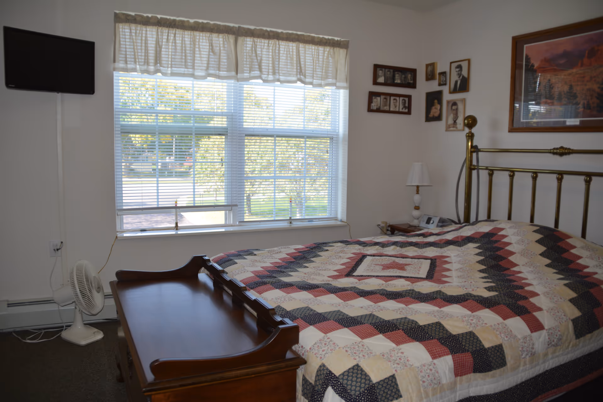 Sunlit bedroom with a brass bed covered by a patchwork quilt, a window with blinds, bedside table and a small wall-mounted TV.