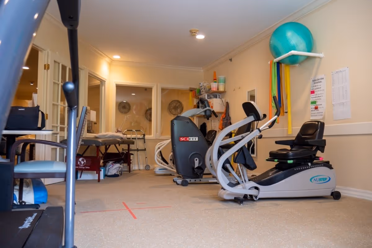 Interior view of a fitness or therapy room in a senior living facility with exercise equipment including a recumbent bike and an elliptical machine. There are colorful resistance bands and a large exercise ball mounted on the wall. The room has beige walls, a carpeted floor, and some chairs and tables in the background.