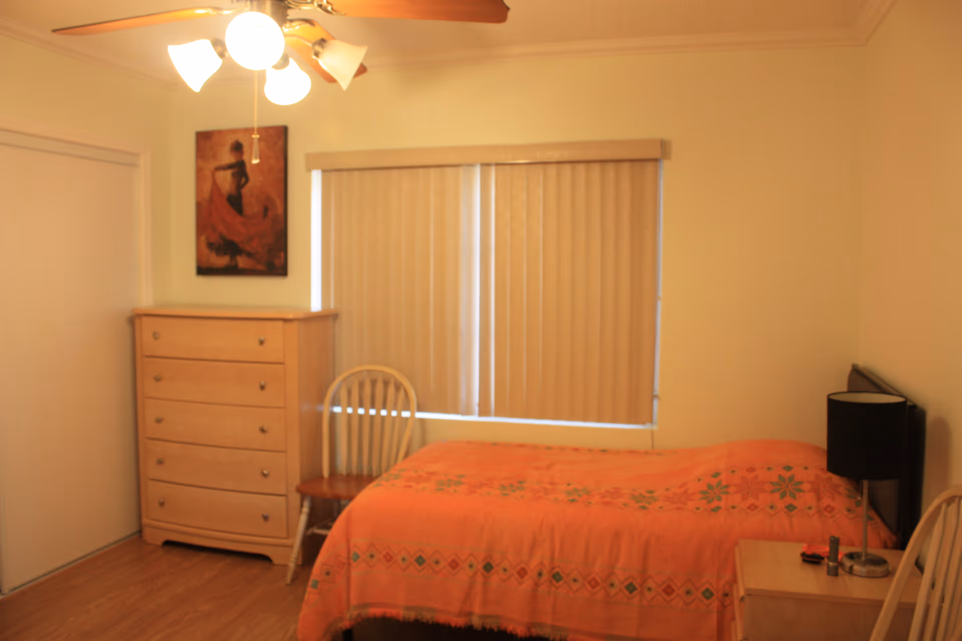 A small bedroom with a dresser, bed, nightstand, chair, blinds, ceiling fan, and peach bedspread.